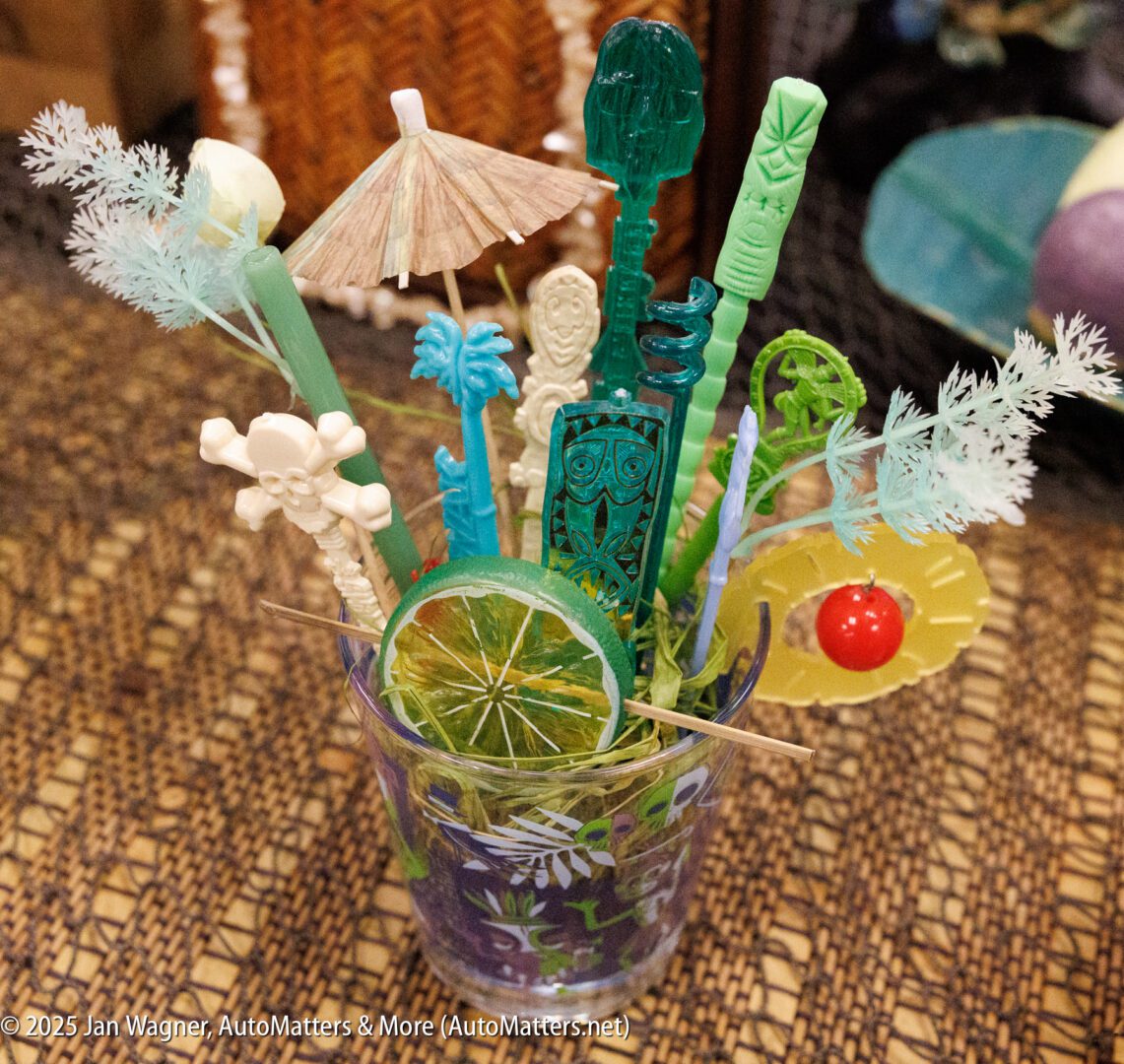 A glass filled with assorted colorful cocktail stirrers, decorative picks, a faux lime slice, and a small paper umbrella on a woven surface.