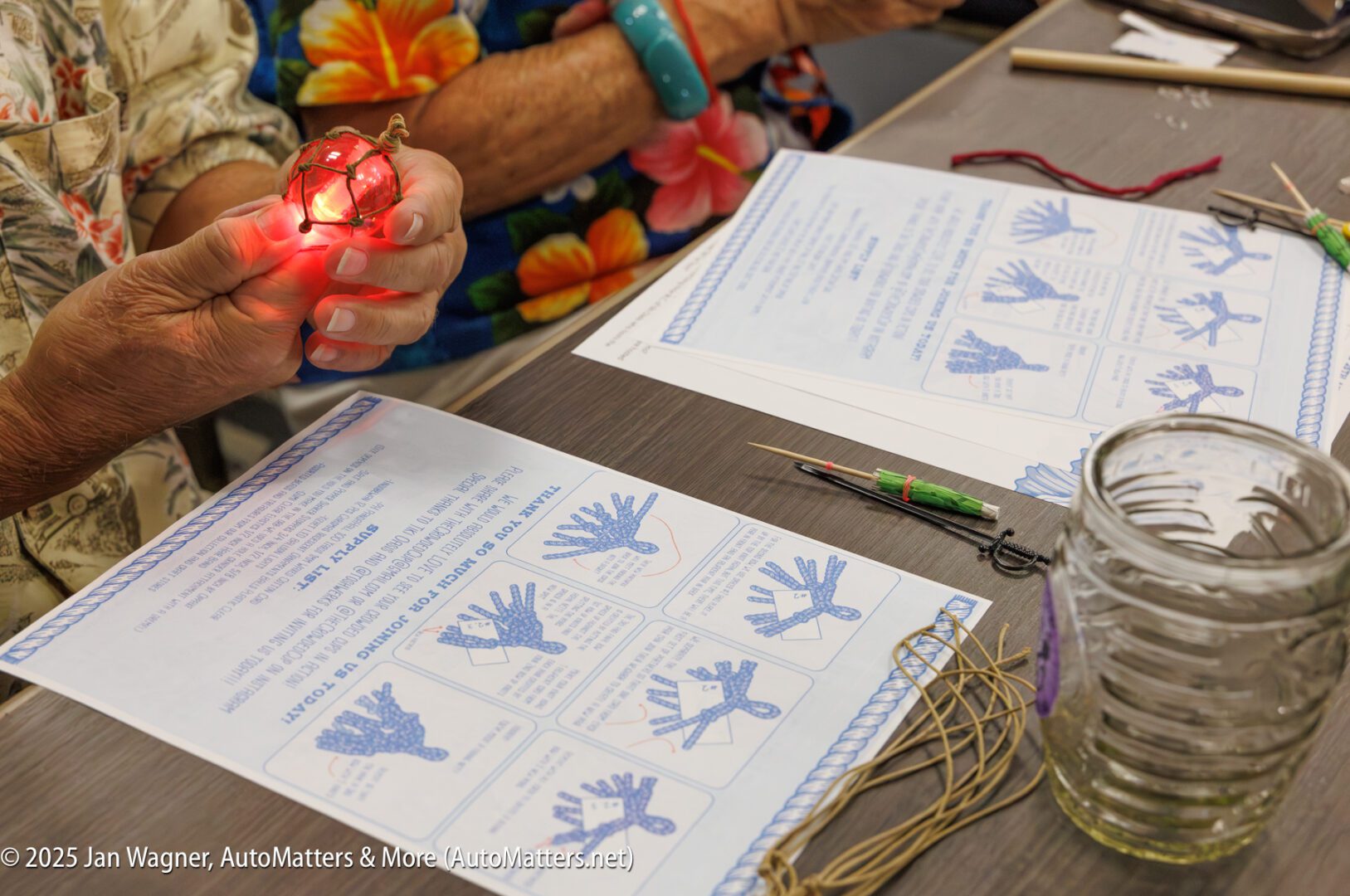 Two people sit at a table working on a craft project. One person holds a red, lit ornament; instructions, string, tools, and a glass jar are on the table.