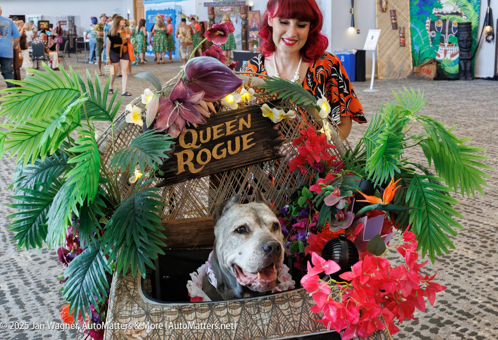 A woman stands behind a cart decorated with tropical plants and flowers, reading "Queen Rogue," with a large dog sitting inside.