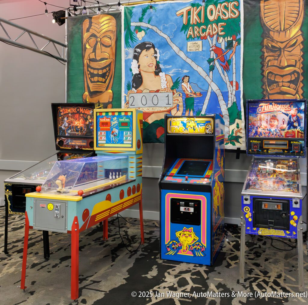 Four vintage arcade and pinball machines are displayed in front of a colorful "Tiki Oasis Arcade" banner with a "2001" sign in a carpeted room.