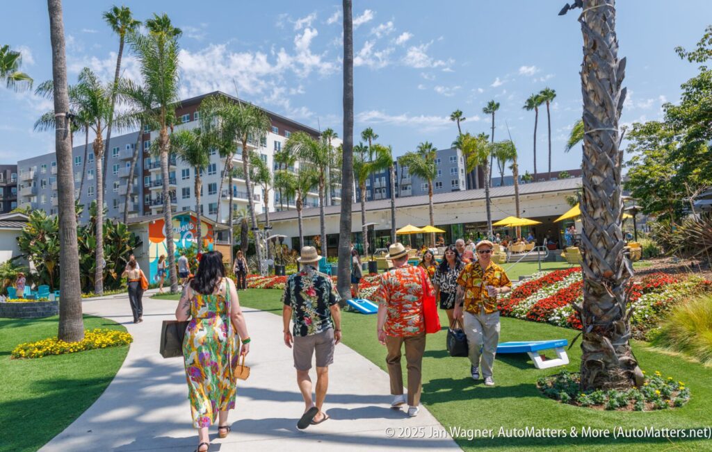 People in colorful clothing walk along a landscaped path with palm trees, flowers, and outdoor seating near modern apartment buildings on a sunny day.