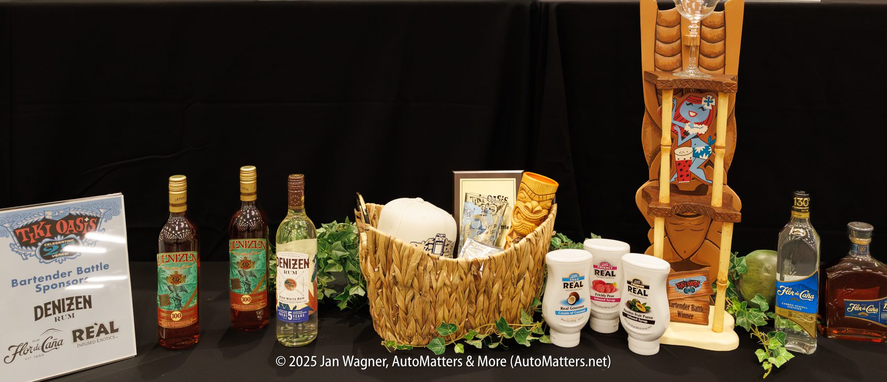 A display of cocktail ingredients, tiki mugs, rum bottles, and branded cups on a black tablecloth at a bartender event.