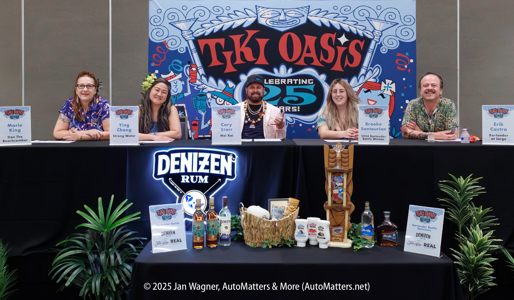 Five panelists sit at a table with Tiki Oasis signs, rum bottles, and plants in front of them. Name cards identify each person. A banner behind them reads “Tiki Oasis Celebrating 25 Years!”.