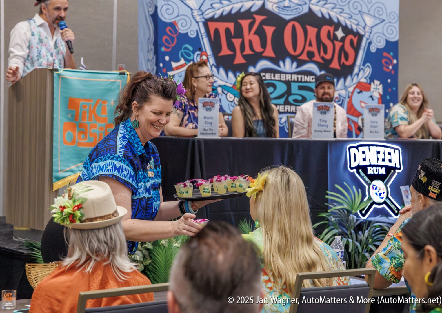 A woman in a blue shirt hands out drinks to seated attendees at a Tiki Oasis event, with a panel of speakers at a table in the background.