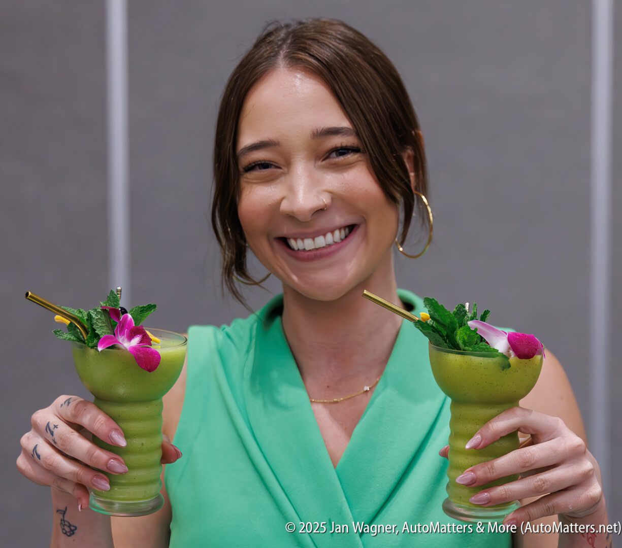 A woman in a green top smiles and holds two green drinks garnished with flowers and mint, each with a straw.