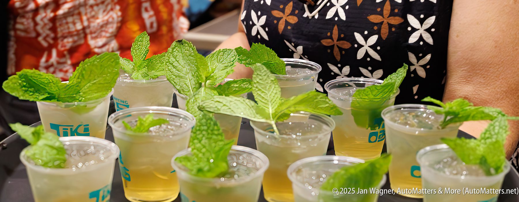 A close-up of several plastic cups filled with a light-colored beverage and garnished with fresh mint leaves, held by people in patterned clothing.