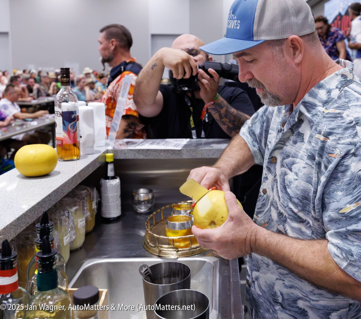 A man in a patterned shirt peels a grapefruit at a bar counter while a crowd, including a photographer, watches in the background. Various bottles and bartending tools are on the counter.