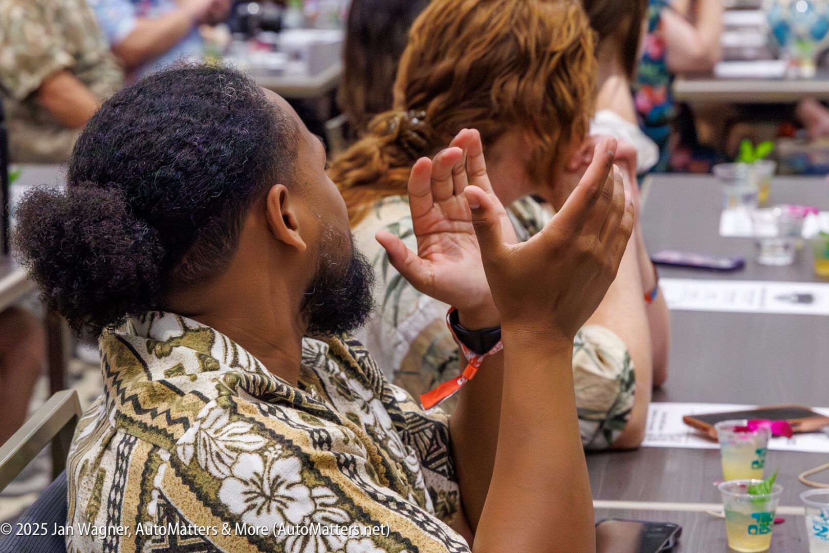 A person in a patterned shirt gestures with their hands while seated at a table among other attendees at an indoor event.