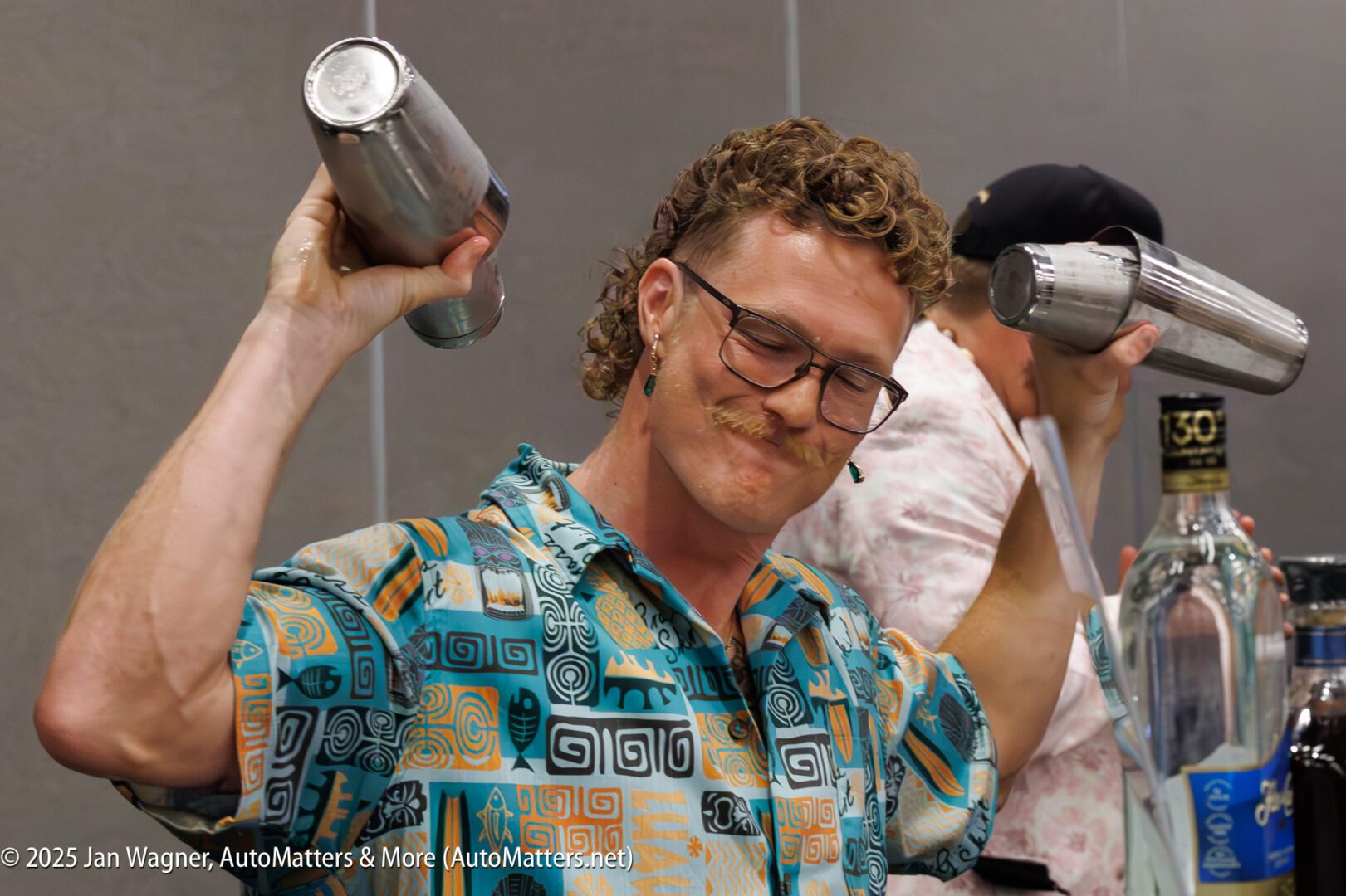 Two bartenders, each holding and shaking cocktail shakers, stand behind a bar with liquor bottles visible, focused on making drinks.
