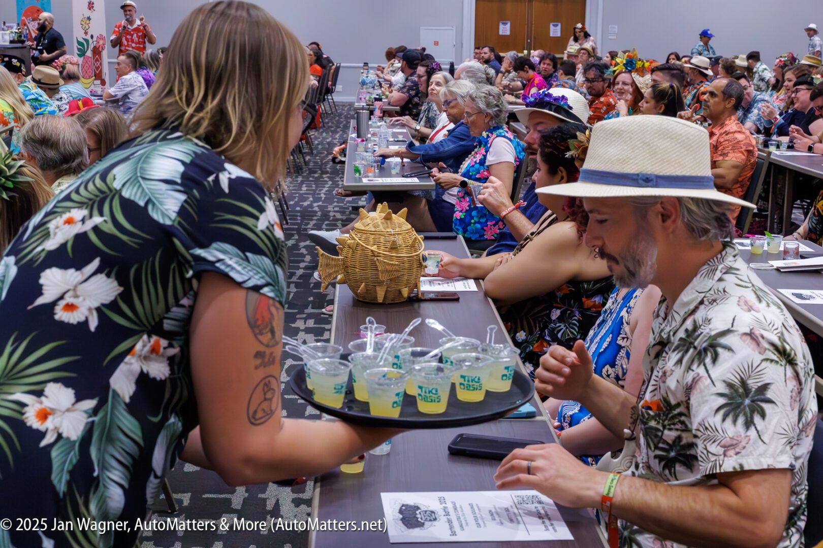 A server hands out drinks to attendees seated at long tables during a tropical-themed event in a conference room.