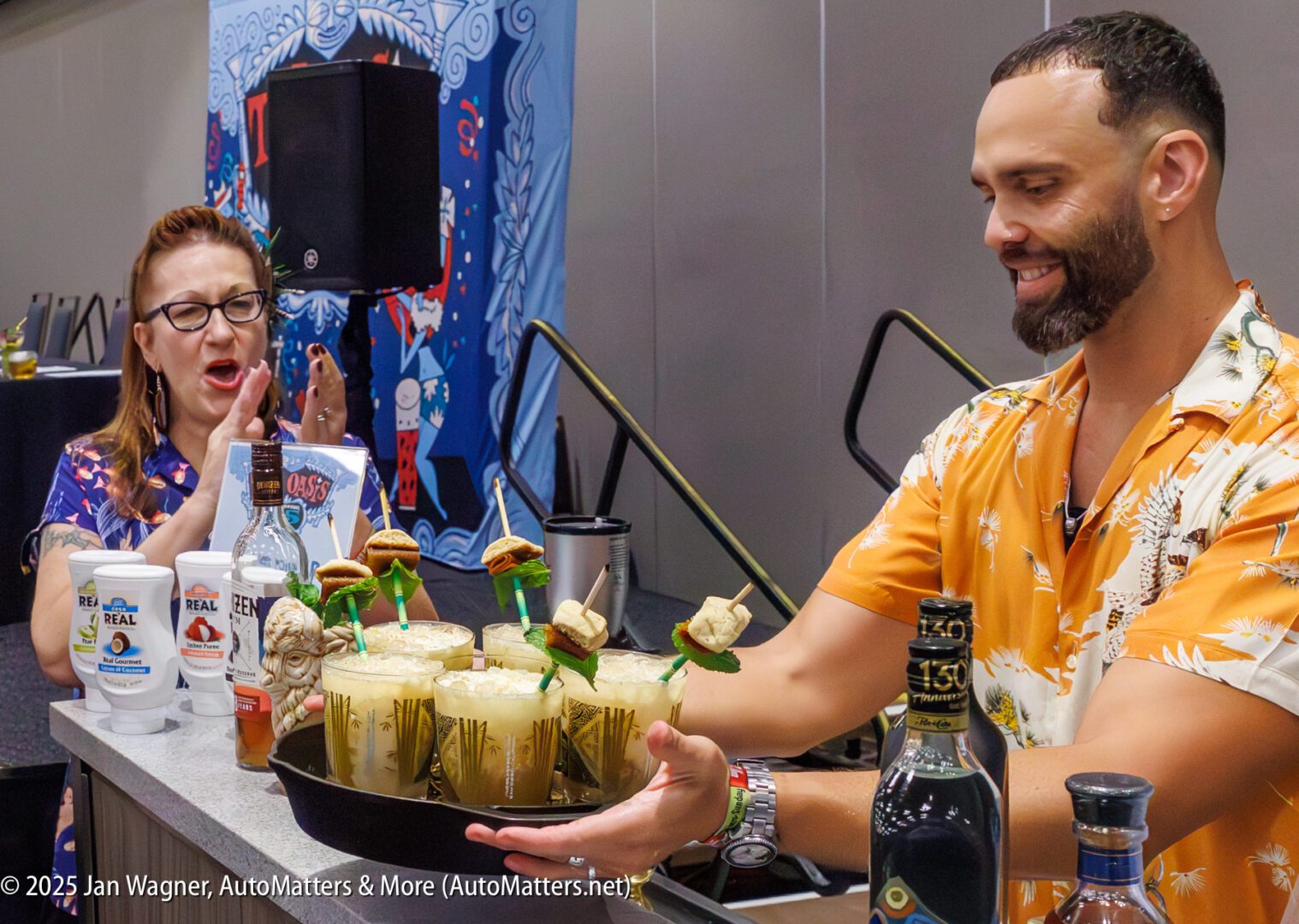 A man in a patterned shirt presents a tray of tropical cocktails garnished with sliders to a woman at a beverage demonstration table.