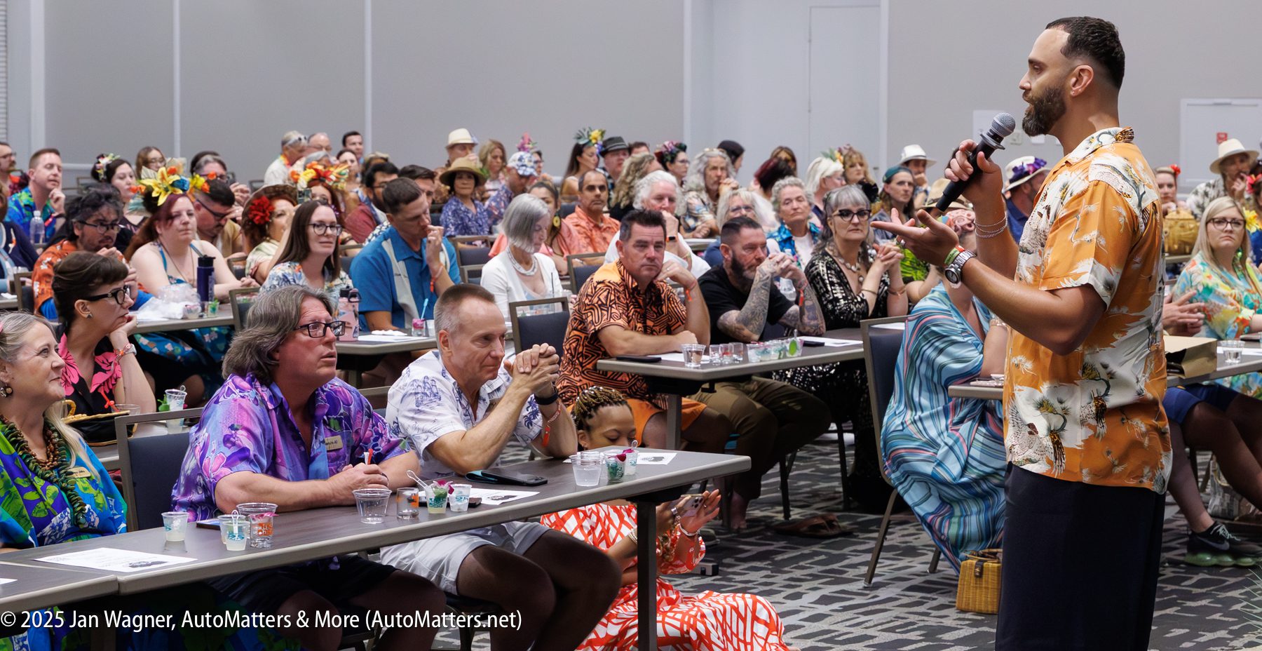 A man in a floral shirt speaks into a microphone to a large seated audience, many of whom are dressed in colorful, tropical-themed clothing.