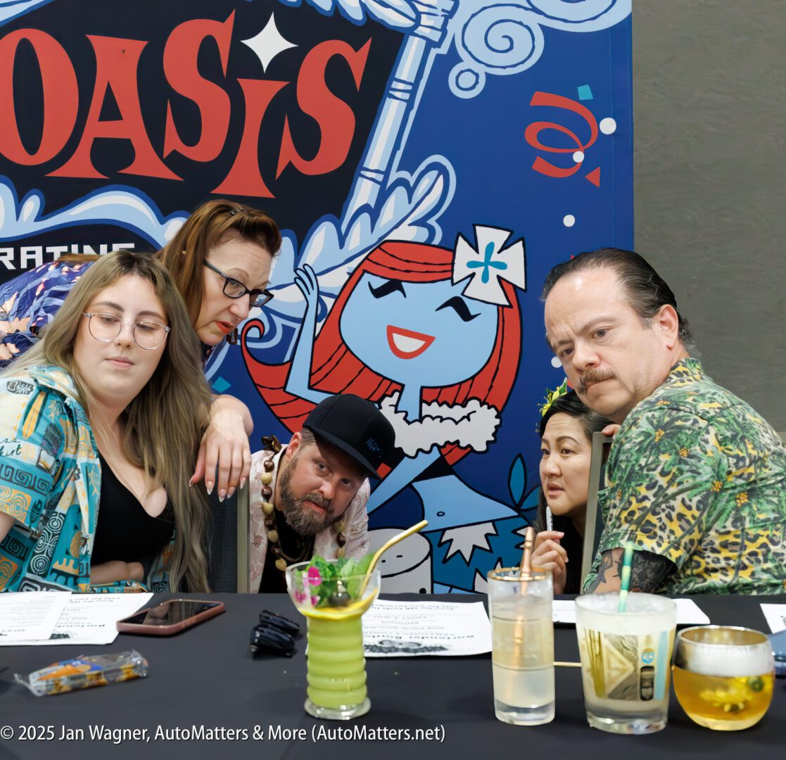 Five adults sit closely together at a table with drinks, posing in front of a colorful Oasis-themed backdrop featuring a cartoon character.