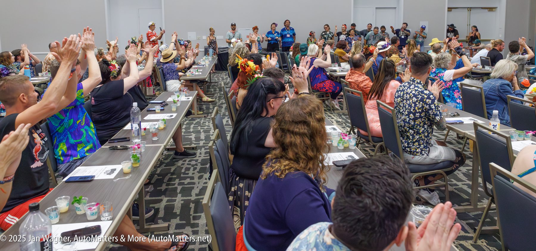 A large group of people in colorful attire sit and raise hands in a conference room, facing speakers at the front. Tables hold papers, drinks, and decorations.
