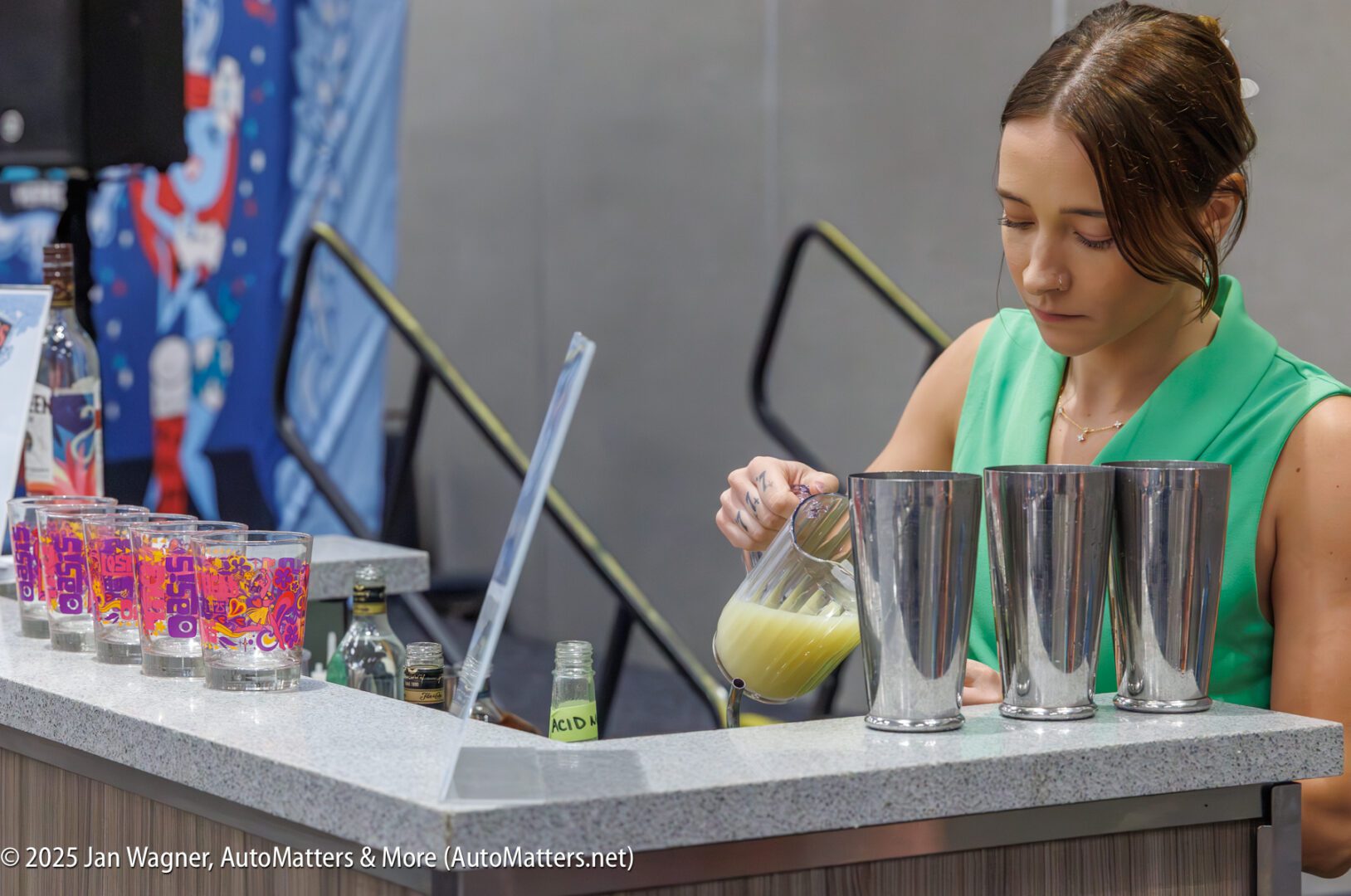 A woman in a green top pours a yellow liquid from a pitcher into a metal tumbler behind a counter with bottles and glasses.