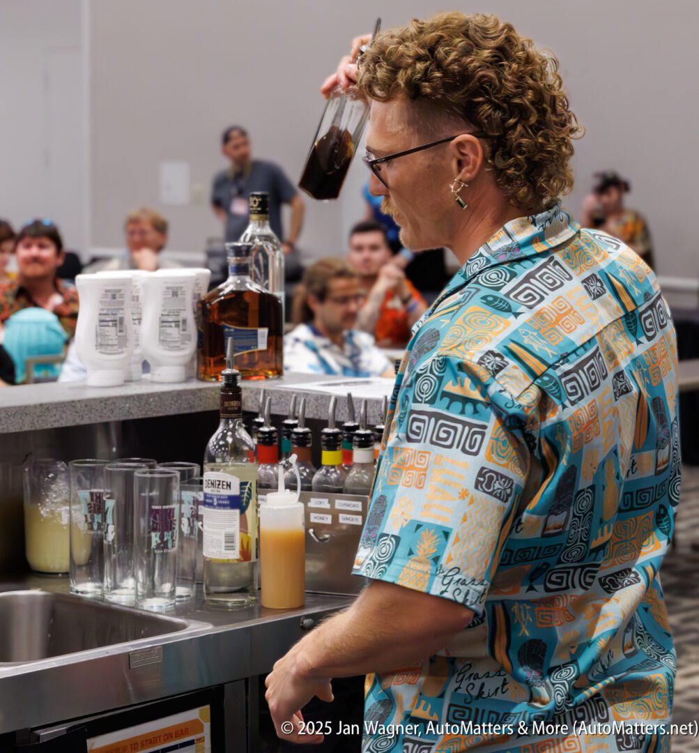 A bartender wearing a patterned shirt prepares drinks at a bar, while a seated audience watches in the background.