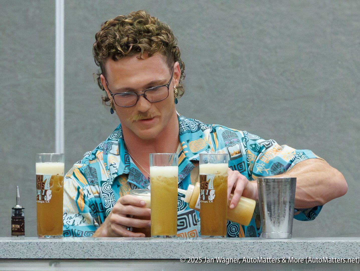 A bartender with curly hair and glasses pours frothy drinks into tall glasses on a bar counter.