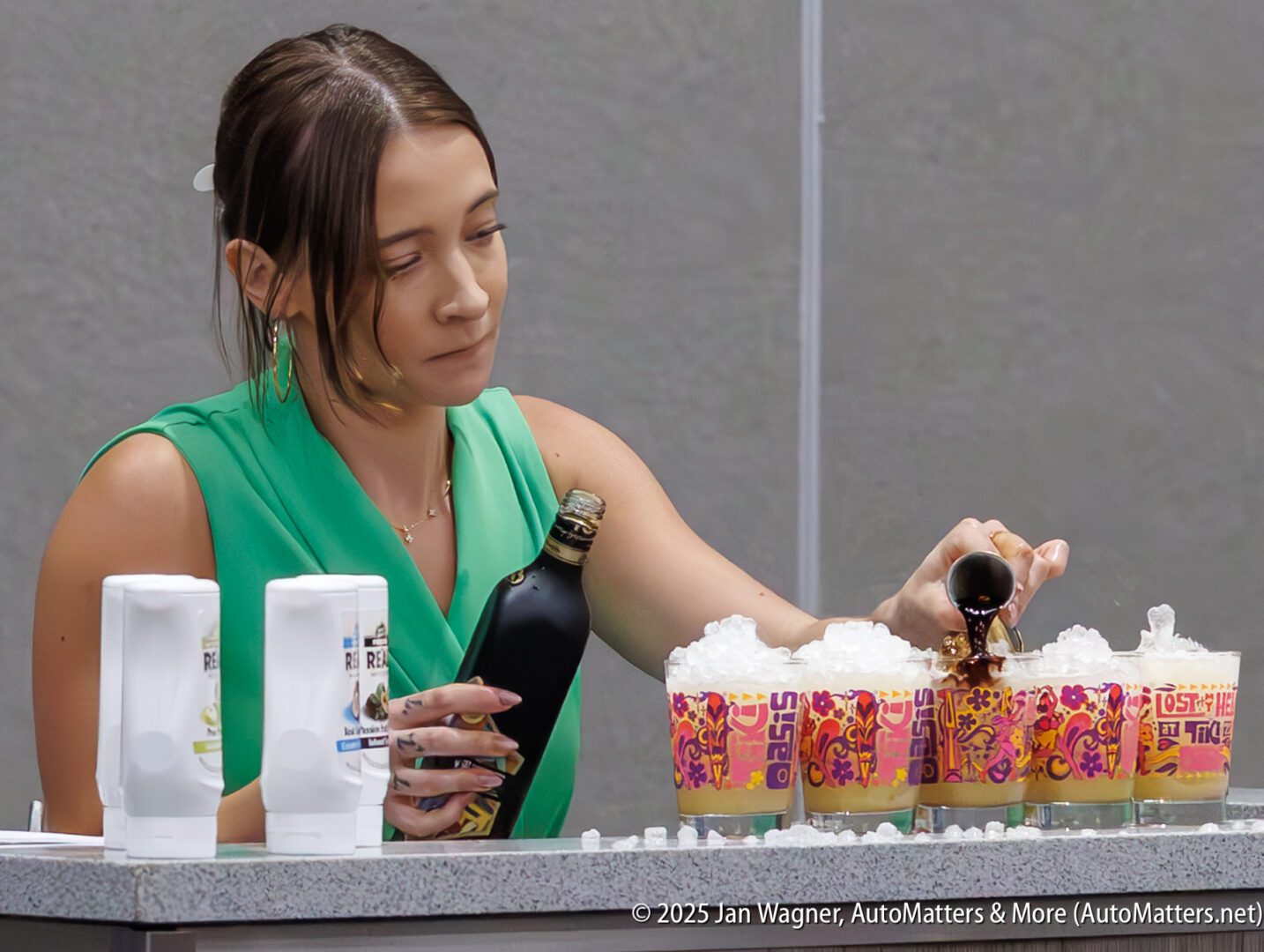 A woman in a green top pours a dark liquid from a bottle into cups filled with ice at a counter, with two hand sanitizer dispensers beside her.