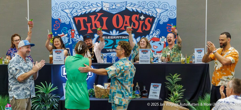 A group of people in tropical attire raise drinks and applaud on stage at a Tiki Oasis event, with banners and decorations in the background.