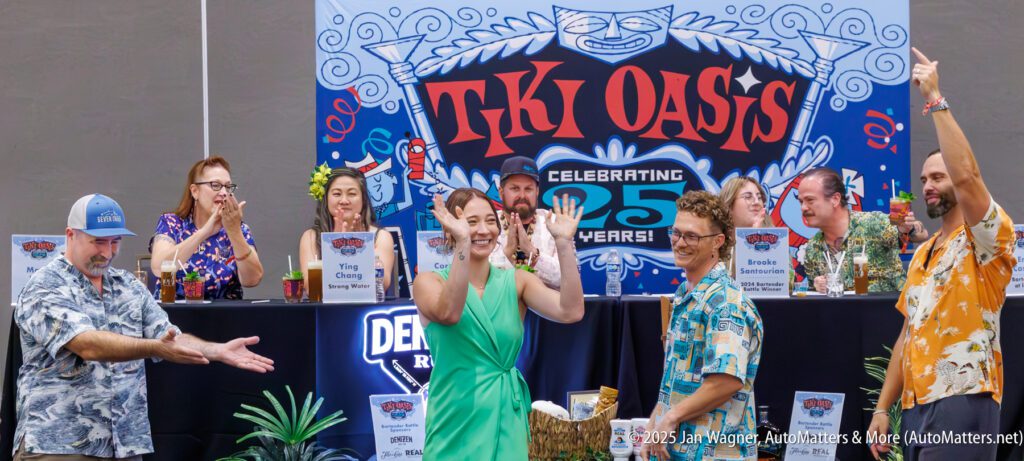 People in tropical attire celebrate on stage at Tiki Oasis 25th anniversary event, with a decorated backdrop and drinks on the table.