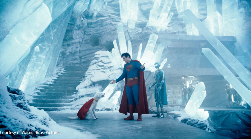 Superman stands in an icy cave with large crystal structures, reaching out to a dog in a red cape. Another figure in metallic armor stands nearby.