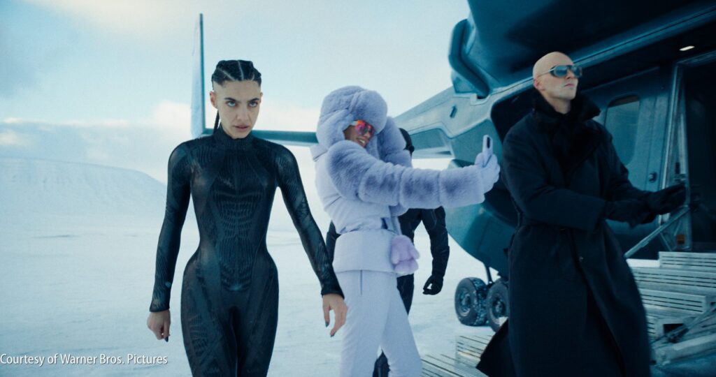 Three people in futuristic outfits walk away from a helicopter in a snowy landscape; one wears a black suit, another a fur coat, and another an all-black coat and sunglasses.