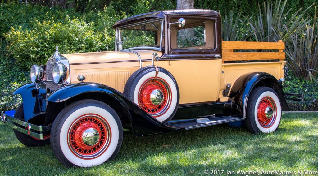 A vintage tan and black pickup truck with red wire wheels and wooden side rails is parked on a grassy lawn.