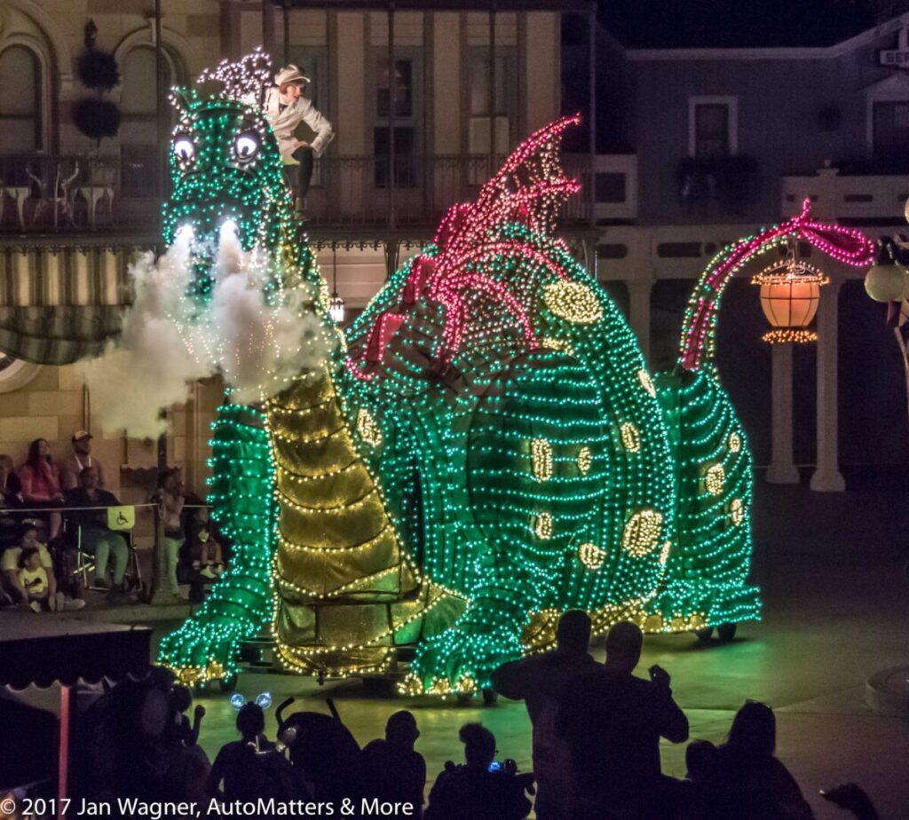A large dragon float covered in green and red lights emits smoke during a nighttime parade, with a performer riding on its head and spectators watching from the sidewalk.