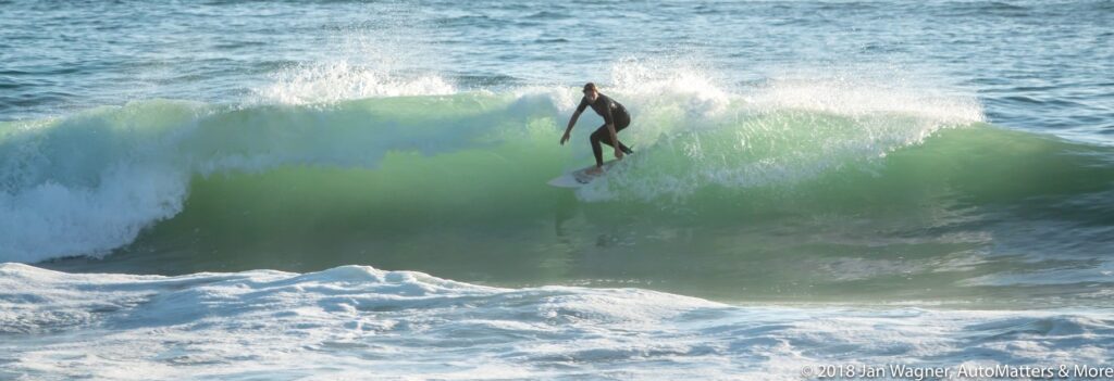 A surfer rides a wave near the shore on a sunny day, with water splashing around and the ocean in the background.
