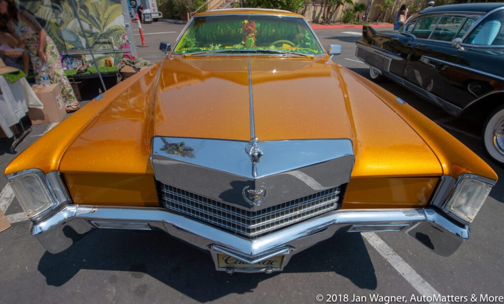 A gold classic Cadillac with a shiny chrome grille is parked at an outdoor car show, with another vintage car partially visible beside it.