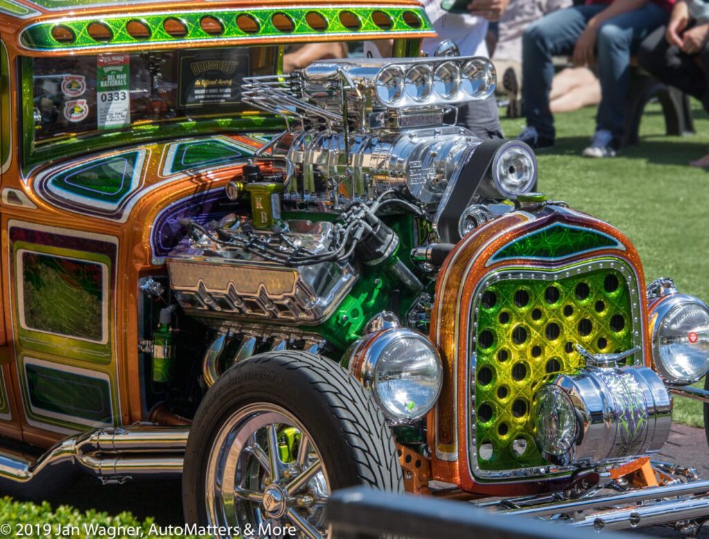 Custom hot rod with a highly detailed, colorful paint job and a large exposed engine on display outdoors, with people in the background.