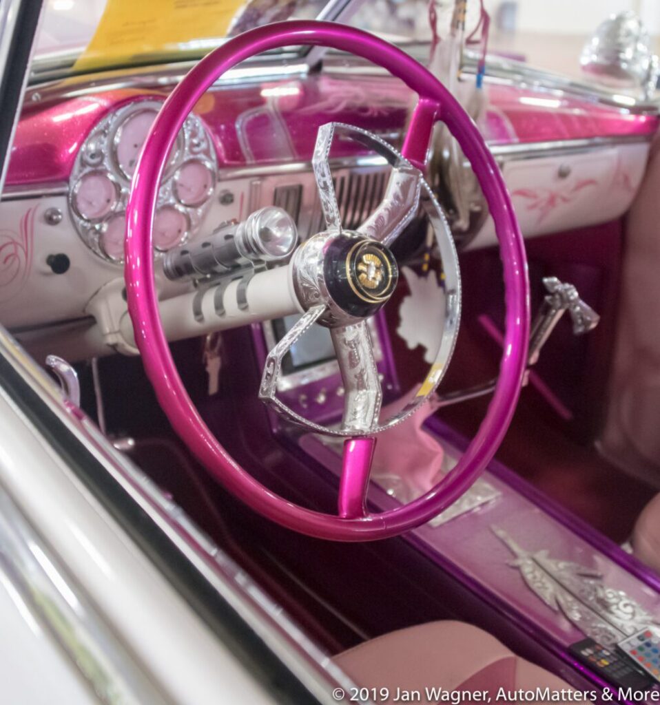 Close-up of a vintage car interior featuring a metallic pink steering wheel, pink dashboard accents, and classic dials.