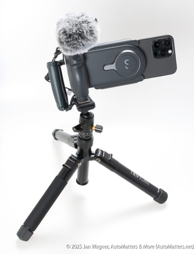 A smartphone mounted horizontally on a mini tripod with a furry microphone attached, set up for recording or streaming.