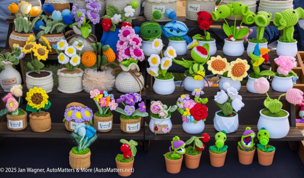 A display of colorful, handmade crocheted flowers and plants arranged in small pots on wooden shelves at a market stall.