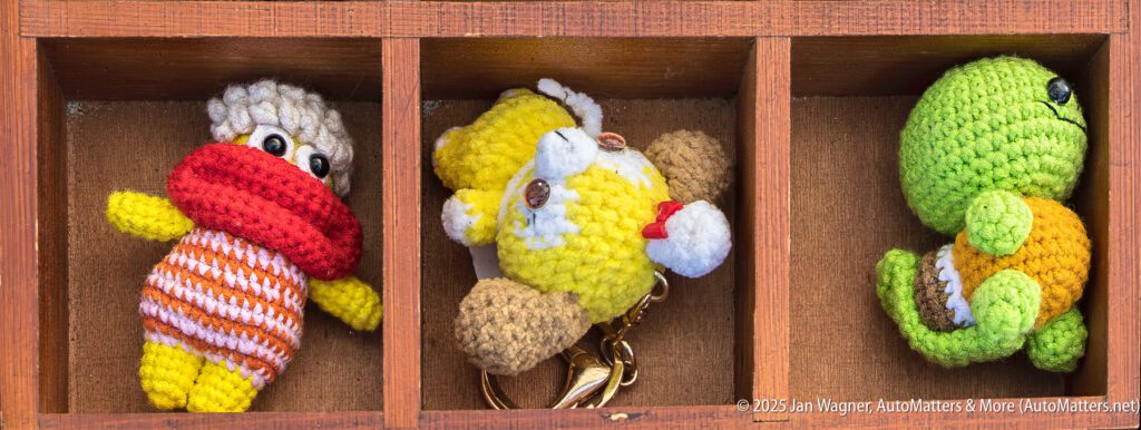 Three small crocheted figurines—a person with a red scarf, a yellow bear keychain, and a green turtle—are displayed in separate wooden compartments.