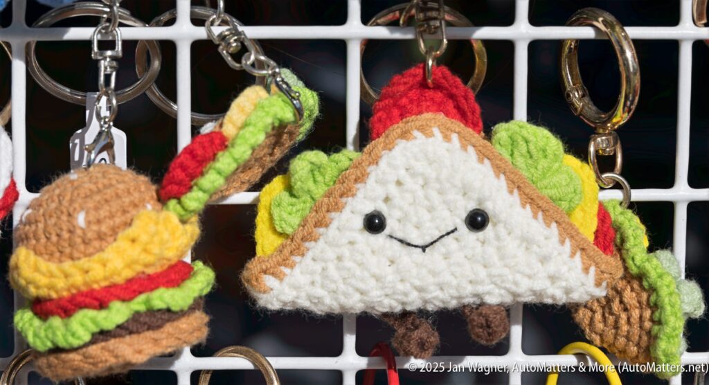 Crocheted keychains shaped like a hamburger and a taco with smiling faces hang on a white display rack.