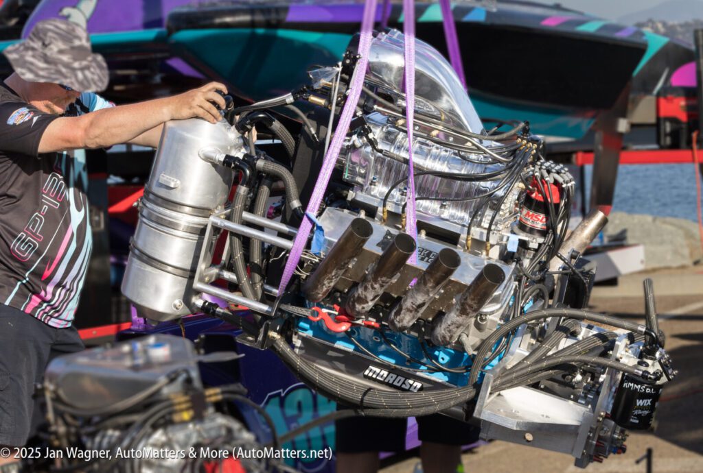 A person guides a large, exposed boat engine being lifted by straps near colorful racing boats at a dock.