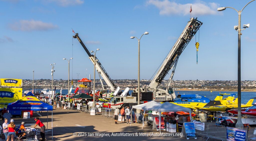 Powerboat event at a marina with cranes, tents, boats, and people gathered along the waterfront under a clear sky.