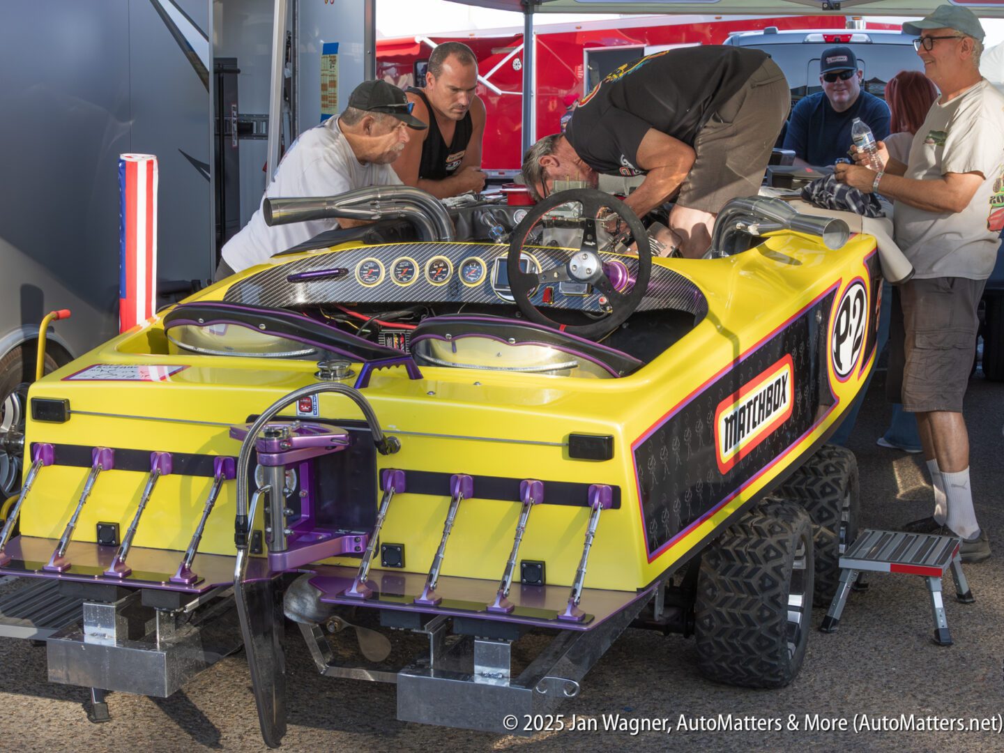 Several men work on a yellow and purple boat with the number 92, which is on a trailer in a paddock area. Tools and equipment are visible around them.