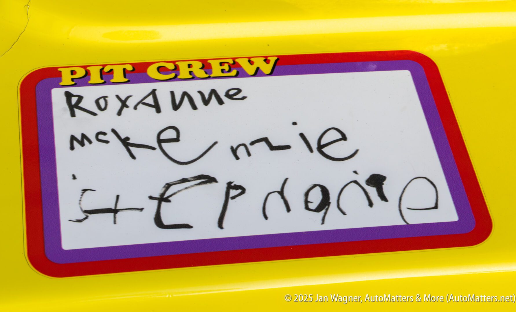 A sticker labeled "PIT CREW" with handwritten names Roxanne, McKenzie, and Stephanie in black marker on a yellow background.