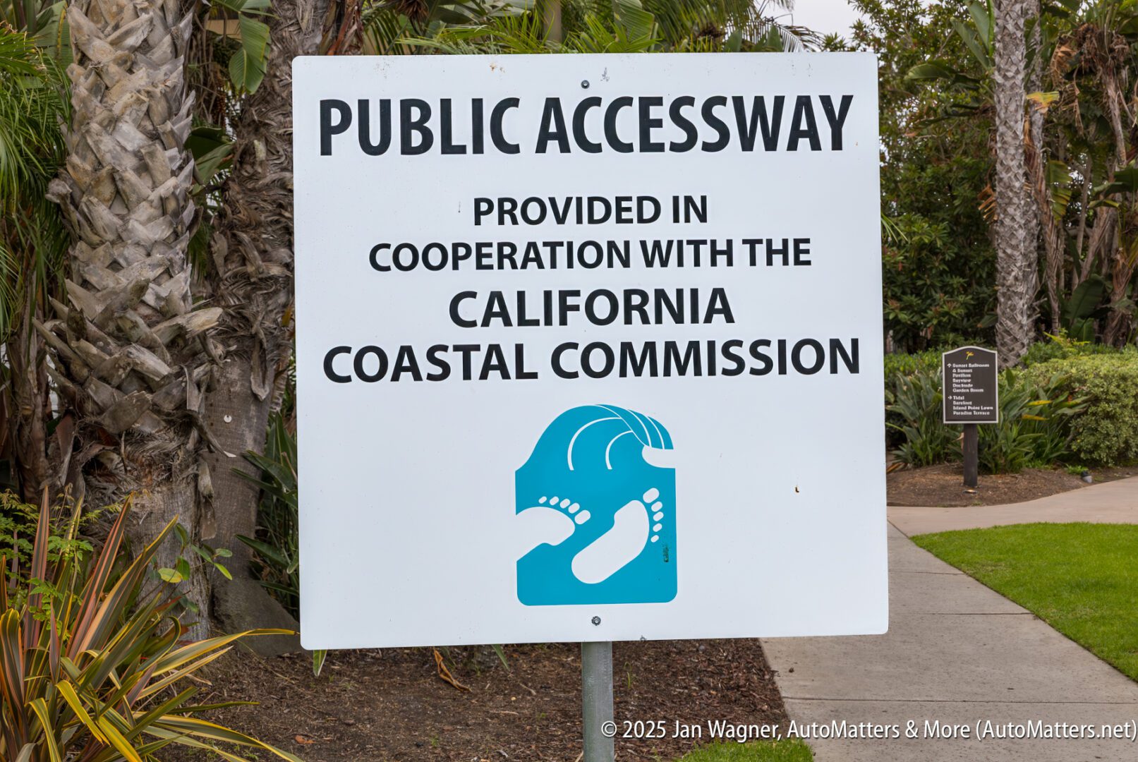 A sign reads "Public Accessway provided in cooperation with the California Coastal Commission" with an icon of two feet and ocean waves, near a landscaped walkway.