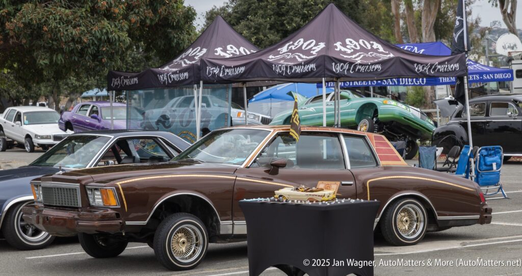 A brown classic car with gold rims is displayed at an outdoor auto show, with other vintage cars and tents visible in the background.