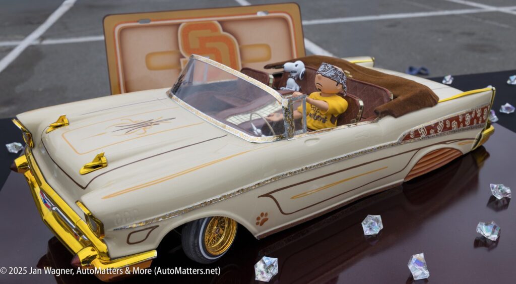 A detailed model of a cream-colored lowrider car with gold accents and a small dog figure in the passenger seat, displayed outdoors with crystal decorations nearby.