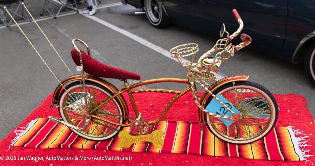 A gold and red custom lowrider bicycle with ornate details is displayed on a red striped blanket at an outdoor event.