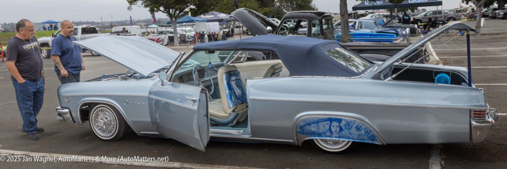 A silver convertible car with a blue soft top is parked at a car show with its doors, trunk, and hood open; two men stand nearby observing.