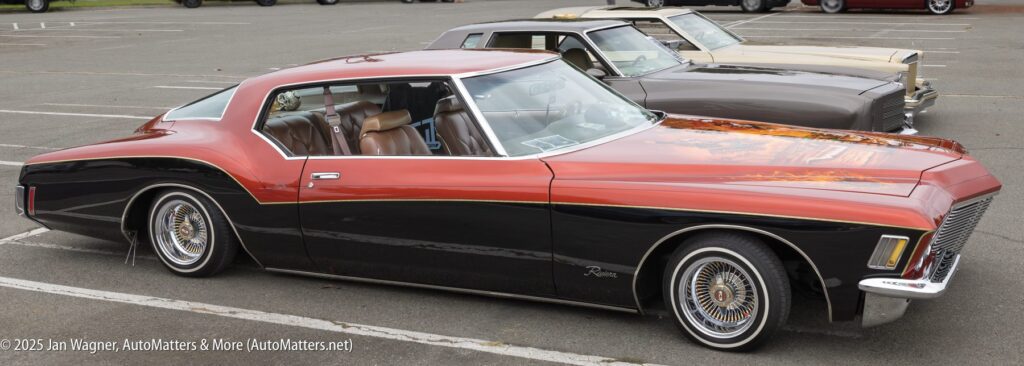 A vintage red and black Buick Riviera car with whitewall tires is parked in a lot next to another classic car.