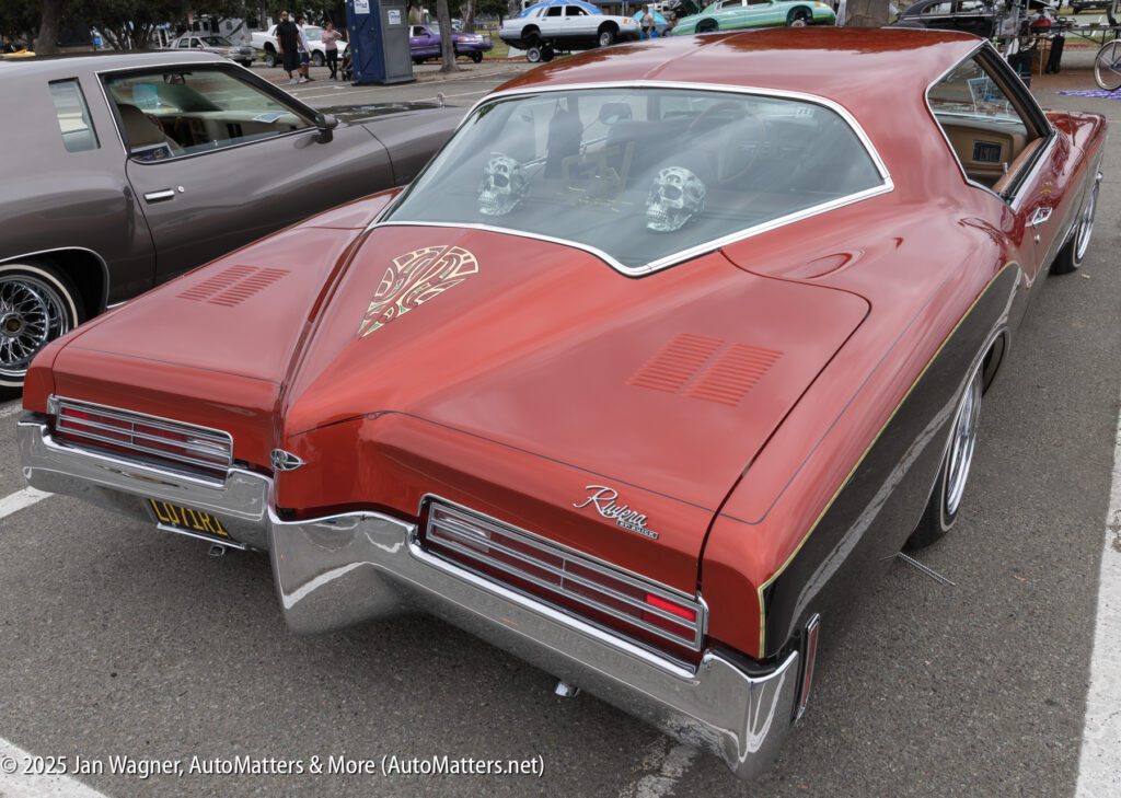 A red Buick Riviera coupe with a split rear window, parked at a car show; custom artwork is visible on the trunk lid.