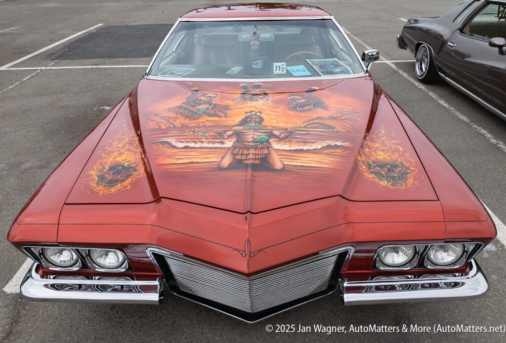 A classic car with detailed mural art of Indigenous figures and landscapes painted on the hood, parked in a lot with another car nearby.
