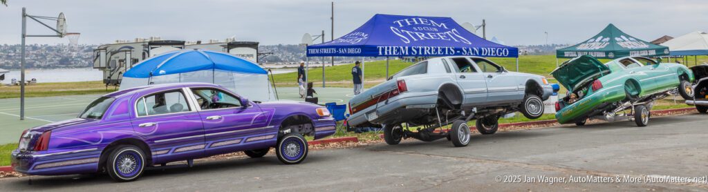 Three customized lowrider cars with colorful paint are parked in a row, two with raised hydraulics, near a park with tents and a basketball court in the background.