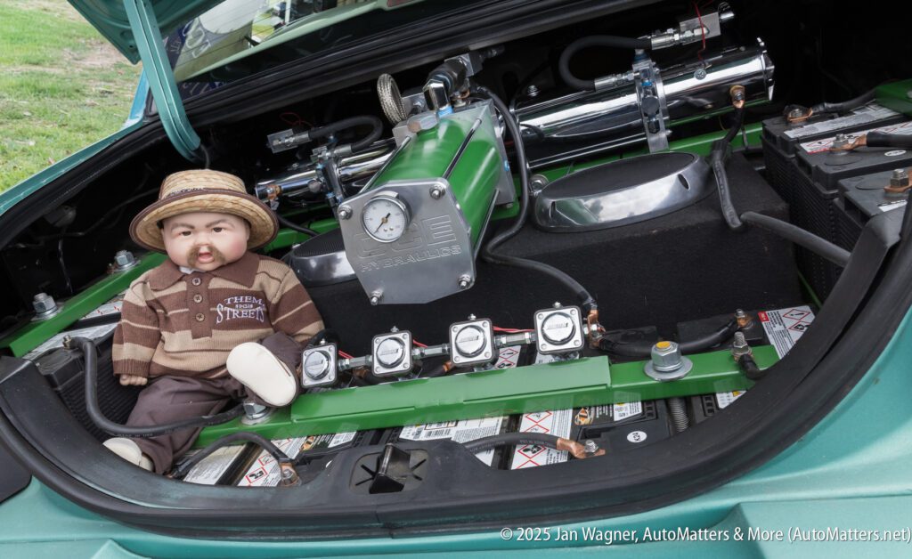 A doll in a striped hat and brown outfit sits inside a car trunk next to exposed mechanical equipment and multiple batteries.
